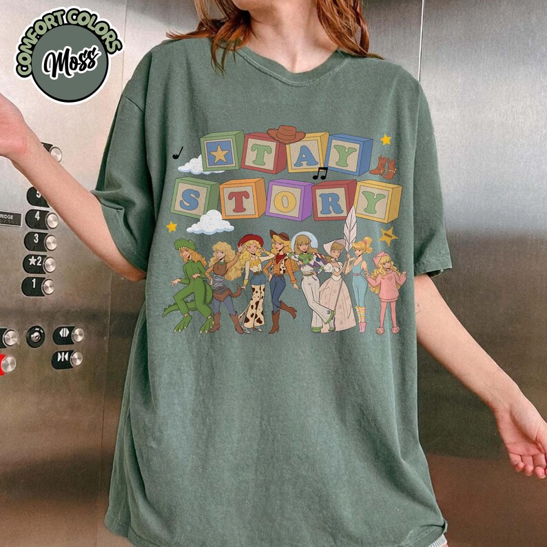 May include: A moss green short-sleeve t-shirt featuring a graphic print of colourful alphabet blocks spelling out "TAY STORY" above a line of illustrated characters dressed in various outfits, including a dinosaur suit and space-themed attire.