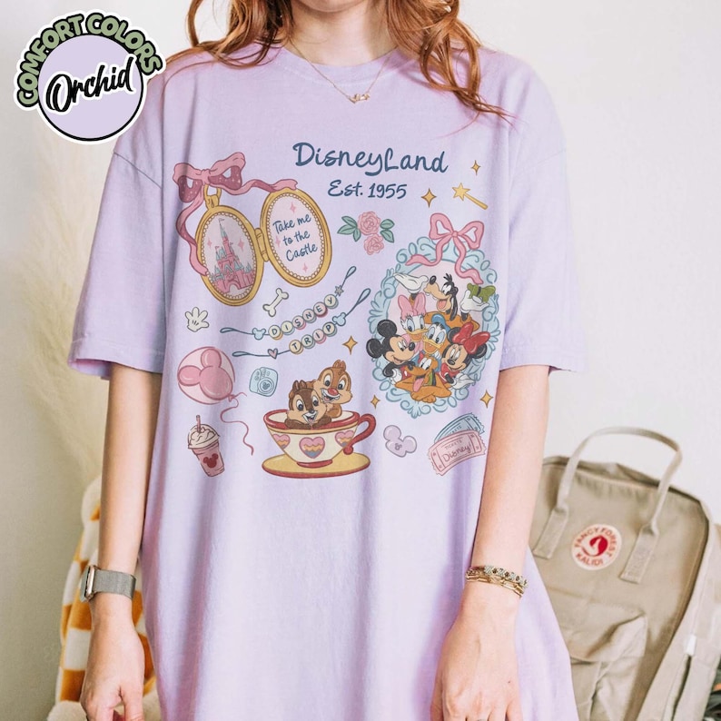 Peut inclure: T-shirt lilas avec des illustrations de Disneyland. Le design comprend Mickey Mouse, Minnie Mouse, Donald Duck et Dingo. D'autres graphiques incluent Tic et Tac dans une tasse &agrave; th&eacute;, un ch&acirc;teau et le texte "Disneyland Est. 1955".