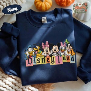 May include: A navy blue crewneck sweatshirt featuring an embroidered graphic of Disney characters including Donald Duck, Pluto, Minnie Mouse, Mickey Mouse, Daisy Duck, and Goofy. Below the characters is the text Disneyland in colorful block letters.