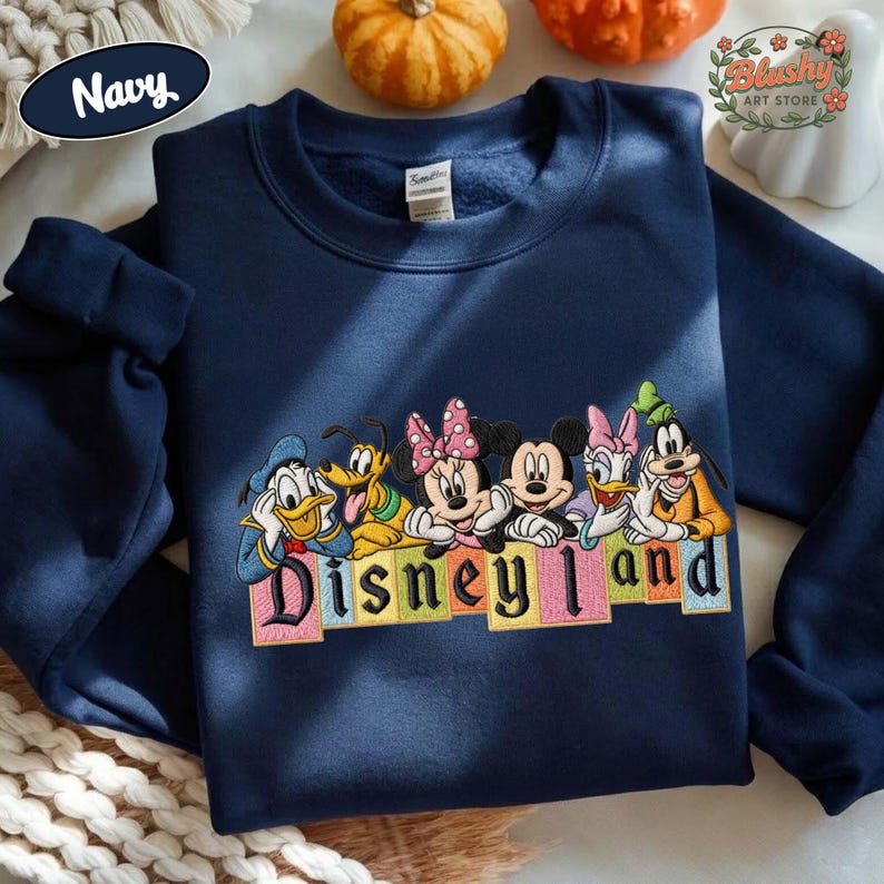 May include: A navy blue crewneck sweatshirt featuring an embroidered graphic of Disney characters including Donald Duck, Pluto, Minnie Mouse, Mickey Mouse, Daisy Duck, and Goofy. Below the characters is the text Disneyland in colorful block letters.