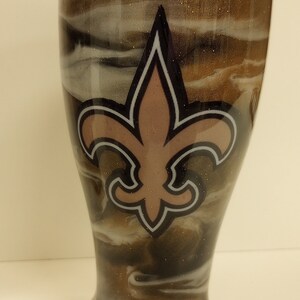 May include: A brown and white swirl tumbler with a gold glitter finish. The tumbler features a black and white fleur-de-lis with a pink centre.