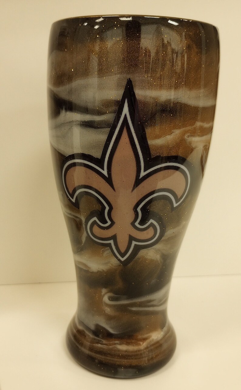 May include: A brown and white swirl tumbler with a gold glitter finish. The tumbler features a black and white fleur-de-lis with a pink centre.