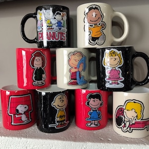 May include: Set of nine ceramic mugs featuring Peanuts characters. The mugs are in various colors, including red, black, white, and cream. Each mug has a different Peanuts character design, including Snoopy, Charlie Brown, Lucy, and Linus. The mugs are all in a similar style, with a rounded shape and a handle.