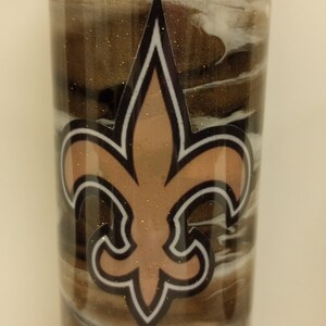 May include: A stainless steel tumbler with a brown, black and white marbled design. The tumbler features a gold fleur-de-lis logo.