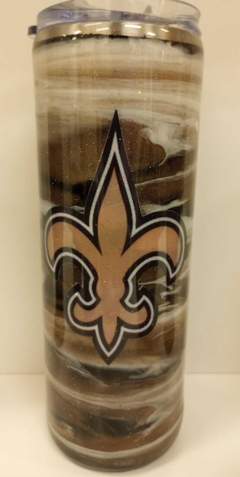 May include: A stainless steel tumbler with a brown, black and white marbled design. The tumbler features a gold fleur-de-lis logo.