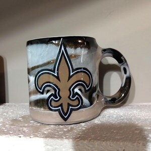 May include: A white ceramic mug with a black handle and a gold and black fleur-de-lis design. The mug has a white, gold, and black marbled background.