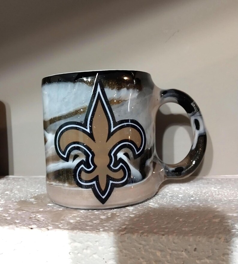 May include: A white ceramic mug with a black handle and a gold and black fleur-de-lis design. The mug has a white, gold, and black marbled background.