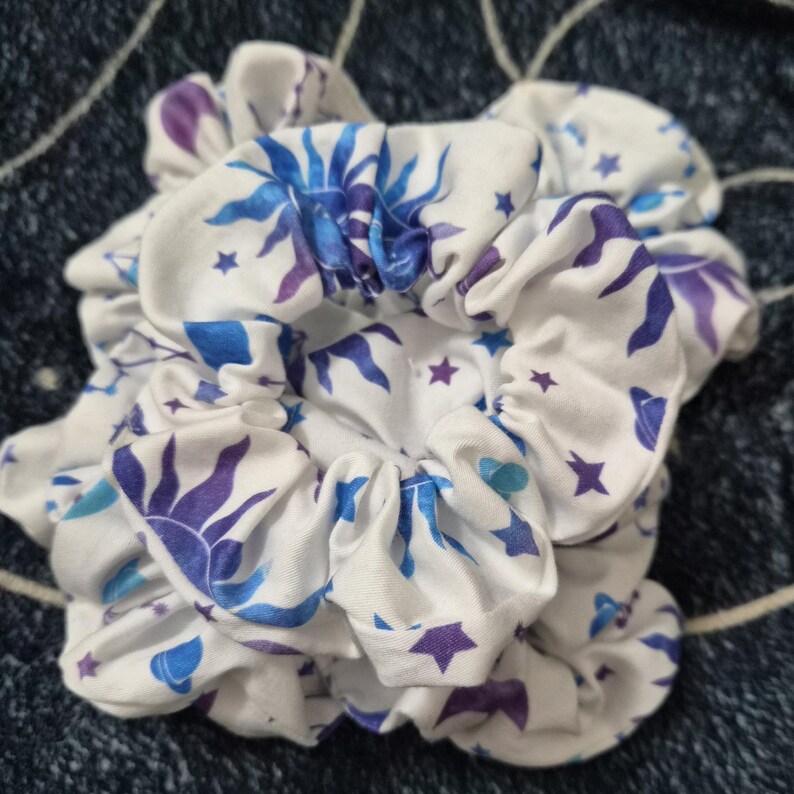 Puede incluir: Un mont&oacute;n de coleteros blancos con un estampado repetido de rayos de sol y estrellas azules y moradas. La tela est&aacute; fruncida y el&aacute;stica, creando una apariencia texturizada. Los coleteros son probablemente accesorios para el cabello.