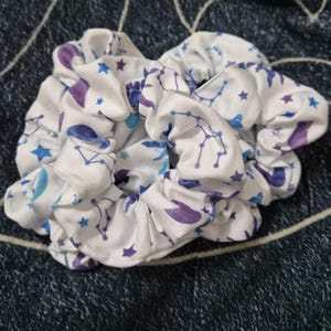 Puede incluir: Coleteros blancos con un estampado celestial con constelaciones, estrellas y lunas crecientes en tonos azules y morados. Estos accesorios para el cabello de tela est&aacute;n fruncidos y rizados.