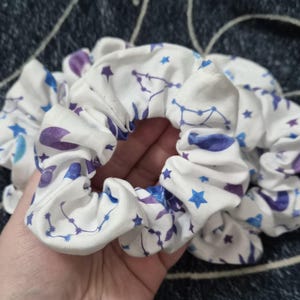May include: White scrunchies with a celestial pattern of blue and purple stars and constellations. The fabric is gathered, creating a ruffled effect. The scrunchies are designed for hair styling.