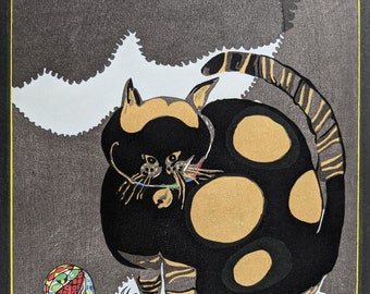 May include: A black and gold cat with large, round spots sits on a white background. The cat has a playful expression and is holding a small, colourful ball in its mouth.