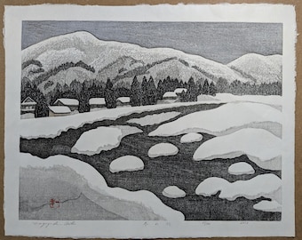May include: A black and white woodblock print depicting a snowy mountain landscape with a river winding through the foreground. The river is partially frozen, with patches of ice and snow visible. A small village is nestled at the base of the mountain, with several houses visible. The print is titled "冬山" in Japanese, which translates to "Winter Mountain".