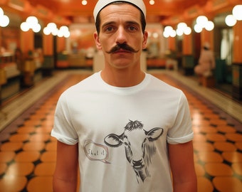 May include: A white t-shirt featuring a black and white illustration of a cow's head with the text "Shut it" in a speech bubble. The person is wearing a white hat.