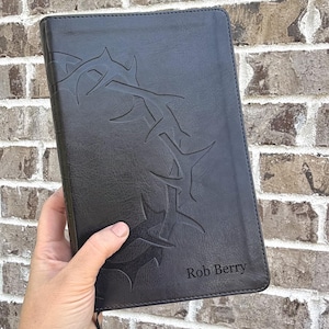 May include: A black rectangular book cover featuring an embossed crown of thorns design on the front. The name Rob Berry is engraved in the bottom right corner. The cover has a smooth texture and a thin black ribbon bookmark attached at the base.