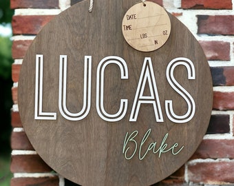 Puede incluir: Un letrero redondo de madera con el nombre "LUCAS" en letras blancas delineadas. Debajo, el nombre "Blake" está escrito en cursiva. Una etiqueta de madera más pequeña cuelga en la parte superior con espacios para fecha, hora, peso en kg y g.