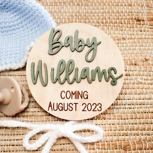 May include: A wooden baby announcement sign with the text "Baby Williams Coming August 2023". The sign is decorated with a blue knitted hat and a beige pacifier.