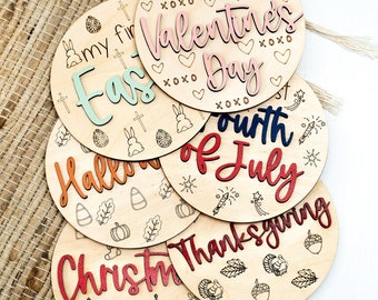 May include: Set of six wooden round ornaments with different holiday themes. Each ornament features a different holiday-themed design and text, including "My First Valentine's Day", "My First Easter", "Halloween", "Fourth of July", "Thanksgiving", and "Christmas".