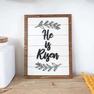 May include: A wooden sign with a white background and a brown frame. The sign says "He is Risen" in black lettering with green leafy sprigs above and below the text.