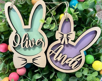May include: Two wooden bunny ornaments with cut-out designs. The bunny on the left is painted green and has the name "Oliver" written inside. The bunny on the right is painted purple and has the name "Amai" written inside.