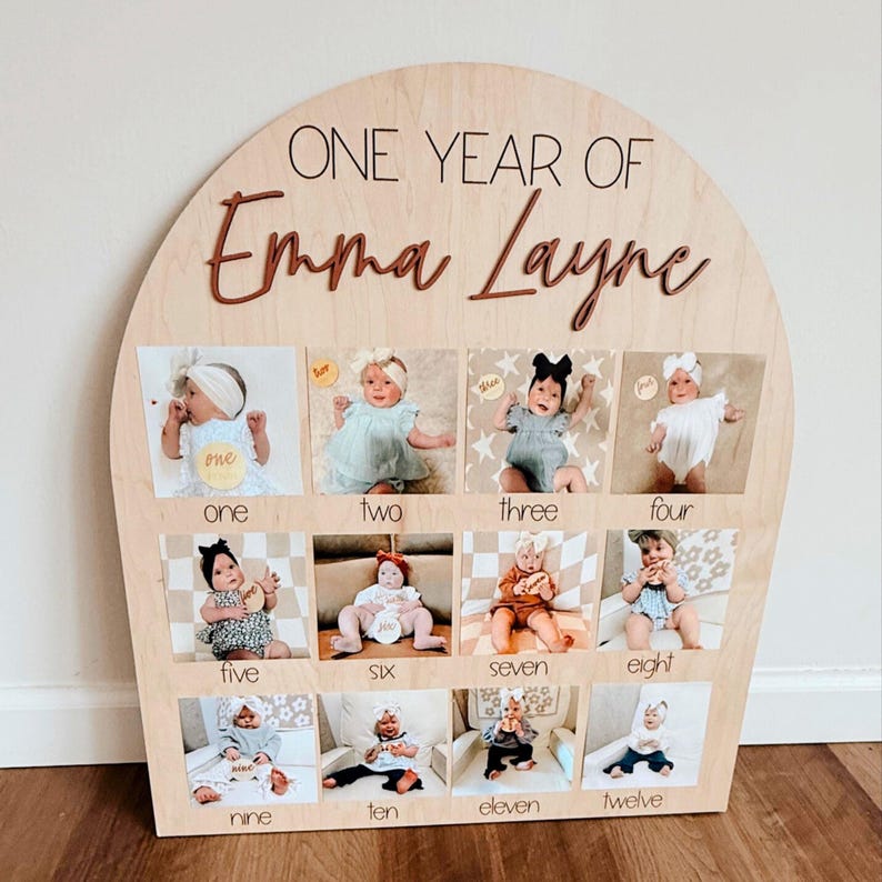 K&ouml;nnte beinhalten: Foto-Display-Board aus Holz mit dem Text "ONE YEAR OF Emma Layne". Das Board hat eine halbkreisf&ouml;rmige Oberseite und enth&auml;lt zw&ouml;lf kleine Fotos, die jeweils mit einem Monat von eins bis zw&ouml;lf beschriftet sind. Die Fotos zeigen ein Baby.