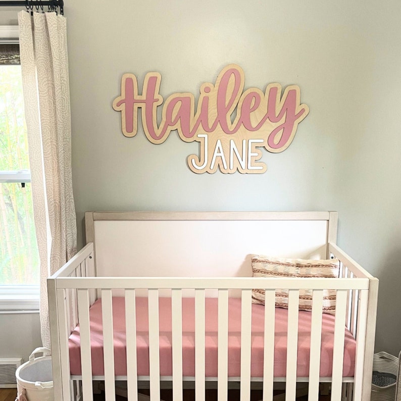 Custom wooden nursery name sign above a  A white crib with a pink sheet and a tan and white patterned pillow. A pink and white wooden sign with the name "Hailey Jane" is on the wall behind the crib.