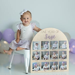 K&ouml;nnte beinhalten: Ein h&ouml;lzernes Foto-Display mit dem Text "ONE YEAR OF Sage" und zw&ouml;lf kleinen Fotoschlitzen. Ein Baby in einem wei&szlig;en Kleid und einer wei&szlig;en Schleife sitzt in der N&auml;he des Displays. Ballons in Lila- und Wei&szlig;t&ouml;nen sind im Hintergrund.