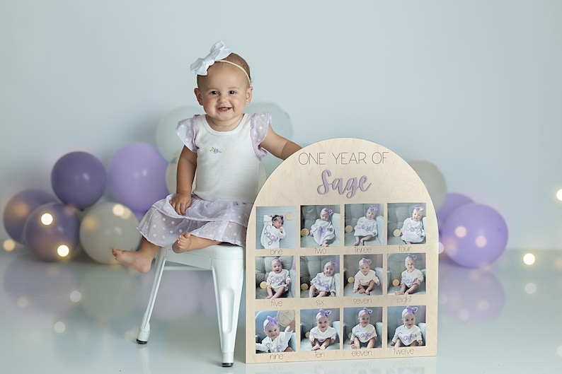 K&ouml;nnte beinhalten: Ein h&ouml;lzernes Foto-Display mit dem Text "ONE YEAR OF Sage" und zw&ouml;lf kleinen Fotoschlitzen. Ein Baby in einem wei&szlig;en Kleid und einer wei&szlig;en Schleife sitzt in der N&auml;he des Displays. Ballons in Lila- und Wei&szlig;t&ouml;nen sind im Hintergrund.