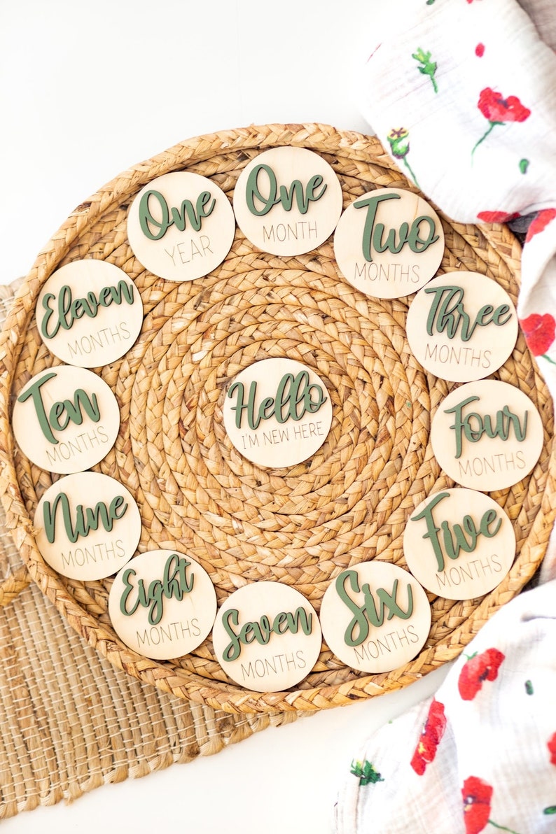 May include: A set of 12 wooden baby milestone discs with green text. The discs feature the words "Hello I'm New Here", "One Year", "One Month", "Two Months", "Three Months", "Four Months", "Five Months", "Six Months", "Seven Months", "Eight Months", "Nine Months", "Ten Months", and "Eleven Months".