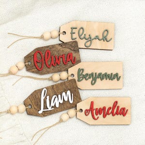 May include: Wooden gift tags with names: Eliyah, Olivia, Liam, Benjamin, and Amelia. Each tag is rectangular with a decorative shape at the top and a string with wooden beads. The names are in various colors: green, red, and white.