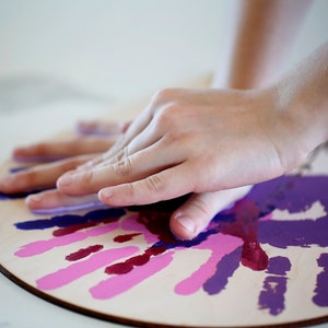 May include: A close-up of a wooden circle with handprints in shades of pink and purple. A hand is pressing down on the circle, leaving a purple handprint.