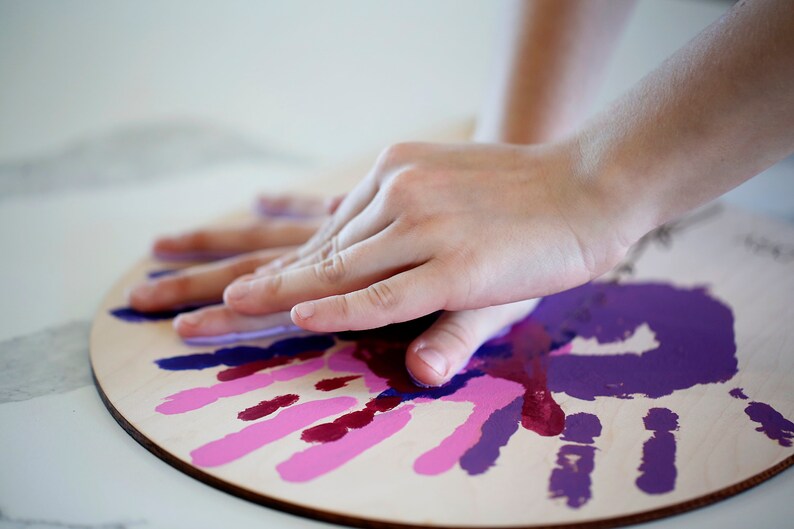 May include: A close-up of a wooden circle with handprints in shades of pink and purple. A hand is pressing down on the circle, leaving a purple handprint.