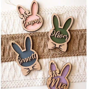 May include: Four wooden Easter bunny decorations, each with a name: Emma (pink), Oliver (green), Bennett (blue), and Mia (purple). Each bunny has a bow and a string. The ornaments are on a white and brown textured surface.
