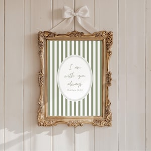 May include: A framed print with a gold ornate frame, featuring a green and white striped background. The print includes the text "I am with you always" and "Matthew 28:20" in a white oval. A white bow is tied at the top.