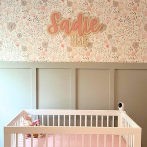 Custom wooden nursery name sign with name "Sadie Mae" in pink and white on a floral wall paper wall above a crib.