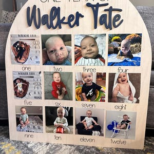 K&ouml;nnte beinhalten: Eine Holz-Fototafel mit dem Text "ONE YEAR OF Walker Tate" in Marineblau. Sie verf&uuml;gt &uuml;ber zw&ouml;lf Fotoschlitze, die jeweils mit einem Monat und einem entsprechenden Foto eines Babys beschriftet sind. Die Tafel ist aus hellem Holz gefertigt.