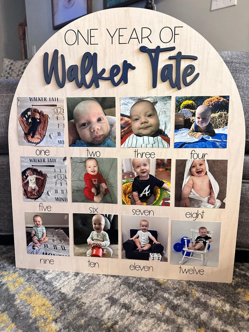 K&ouml;nnte beinhalten: Eine Holz-Fototafel mit dem Text "ONE YEAR OF Walker Tate" in Marineblau. Sie verf&uuml;gt &uuml;ber zw&ouml;lf Fotoschlitze, die jeweils mit einem Monat und einem entsprechenden Foto eines Babys beschriftet sind. Die Tafel ist aus hellem Holz gefertigt.