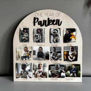 K&ouml;nnte beinhalten: Ein h&ouml;lzerner Foto-Display mit gew&ouml;lbtem Oberteil, mit zw&ouml;lf kleinen Fotoschlitzen, die in einem Raster angeordnet sind. Die Anzeige ist mit "ONE YEAR OF Parker" beschriftet, wobei die Monate unter jedem Foto beschriftet sind. Jedes Foto enth&auml;lt ein Baby.
