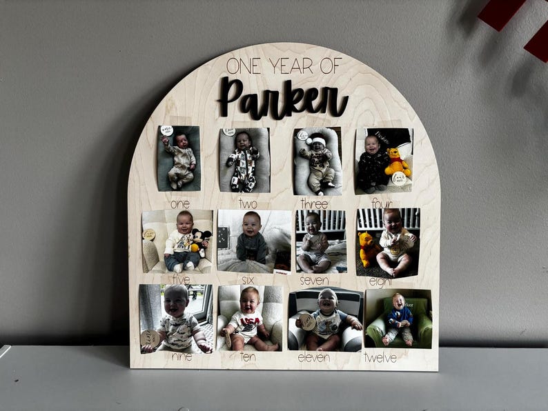 K&ouml;nnte beinhalten: Ein h&ouml;lzerner Foto-Display mit gew&ouml;lbtem Oberteil, mit zw&ouml;lf kleinen Fotoschlitzen, die in einem Raster angeordnet sind. Die Anzeige ist mit "ONE YEAR OF Parker" beschriftet, wobei die Monate unter jedem Foto beschriftet sind. Jedes Foto enth&auml;lt ein Baby.
