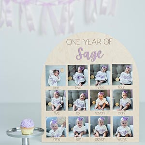 K&ouml;nnte beinhalten: Holz-Fotodisplay mit dem Text "ONE YEAR OF Sage" und zw&ouml;lf Fotoschlitzen. Jeder Schlitz enth&auml;lt ein Babyfoto. Ein Cupcake auf einem silbernen St&auml;nder und ein Lavendel-Banner sind ebenfalls im Bild.