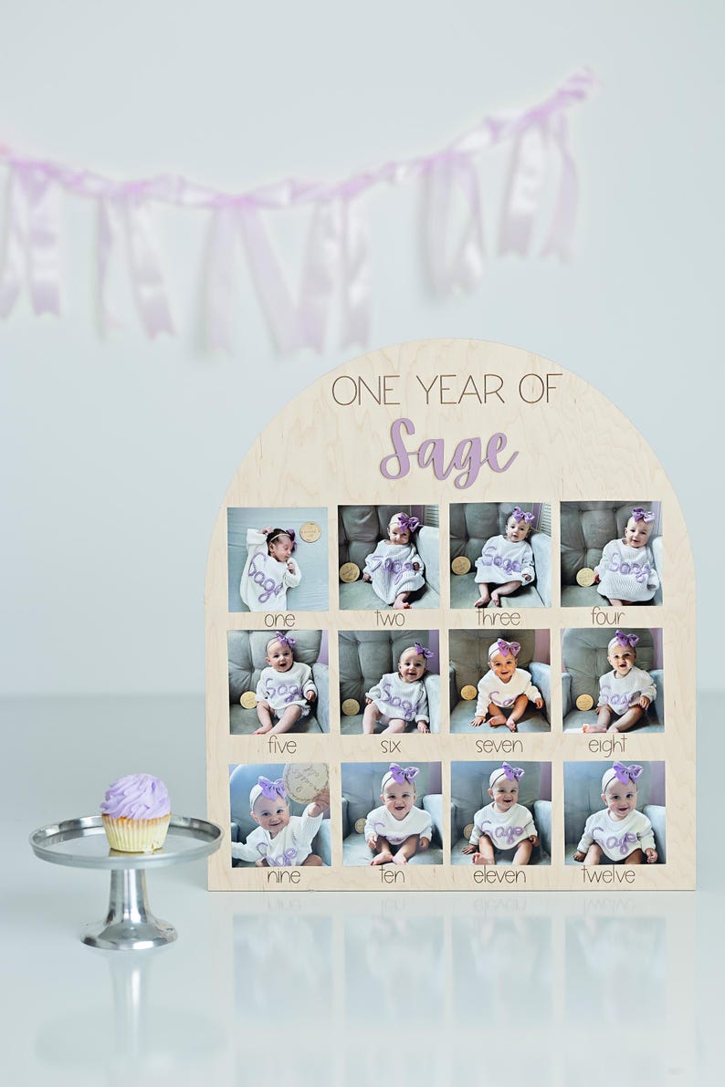 K&ouml;nnte beinhalten: Holz-Fotodisplay mit dem Text "ONE YEAR OF Sage" und zw&ouml;lf Fotoschlitzen. Jeder Schlitz enth&auml;lt ein Babyfoto. Ein Cupcake auf einem silbernen St&auml;nder und ein Lavendel-Banner sind ebenfalls im Bild.