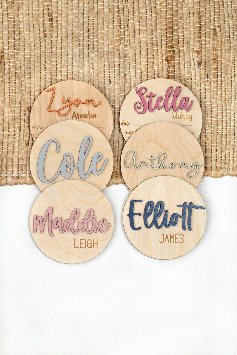 May include: Five wooden round plaques with names written in different colors. The names are:  'Zyon Amelia', 'Cole', 'Stella Makay', 'Anthony', and 'Elliott James'.