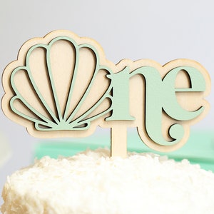 May include: Wooden cake topper with a green seashell and the word "one" in green letters. The topper is perfect for a first birthday party.