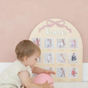 May include: A wooden monthly milestone board with a pink bow and the name 'Rowen' in white script. The board has twelve photo slots, each labeled with a month. A baby in a light-colored outfit plays with a pink balloon near the board.
