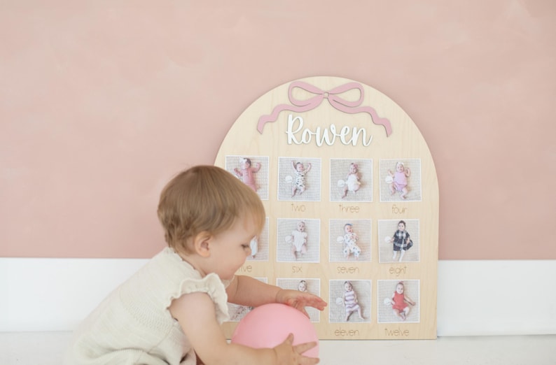 May include: A wooden monthly milestone board with a pink bow and the name 'Rowen' in white script. The board has twelve photo slots, each labeled with a month. A baby in a light-colored outfit plays with a pink balloon near the board.