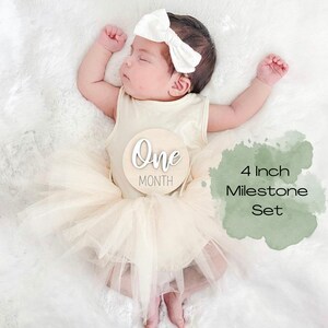 May include: A baby wearing a white headband and a beige tulle skirt is holding a wooden milestone sign that says "One Month". The sign is 4 inches in diameter and is part of a set of milestone signs.
