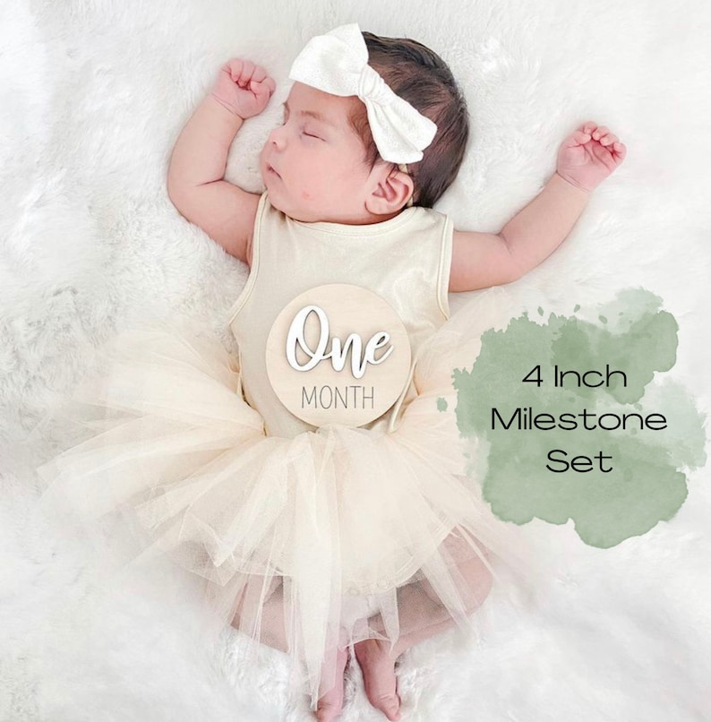 May include: A baby wearing a white headband and a beige tulle skirt is holding a wooden milestone sign that says "One Month". The sign is 4 inches in diameter and is part of a set of milestone signs.