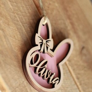 May include: Wooden bunny-shaped ornament with a pink interior and the name "Olivia" in cursive. The ornament has a bow detail and is hanging by a string. The background is a wooden surface.