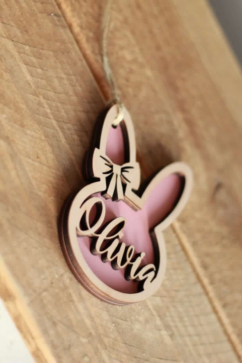 May include: Wooden bunny-shaped ornament with a pink interior and the name "Olivia" in cursive. The ornament has a bow detail and is hanging by a string. The background is a wooden surface.