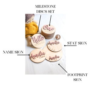 May include: Wooden baby milestone discs set with pink lettering. The set includes a "Hello I'm New Here" disc, a "One Month" disc, and three name discs that say "Amelia Claire". There is also a separate wooden stat sign with spaces for baby's name, date, time, weight, and length.