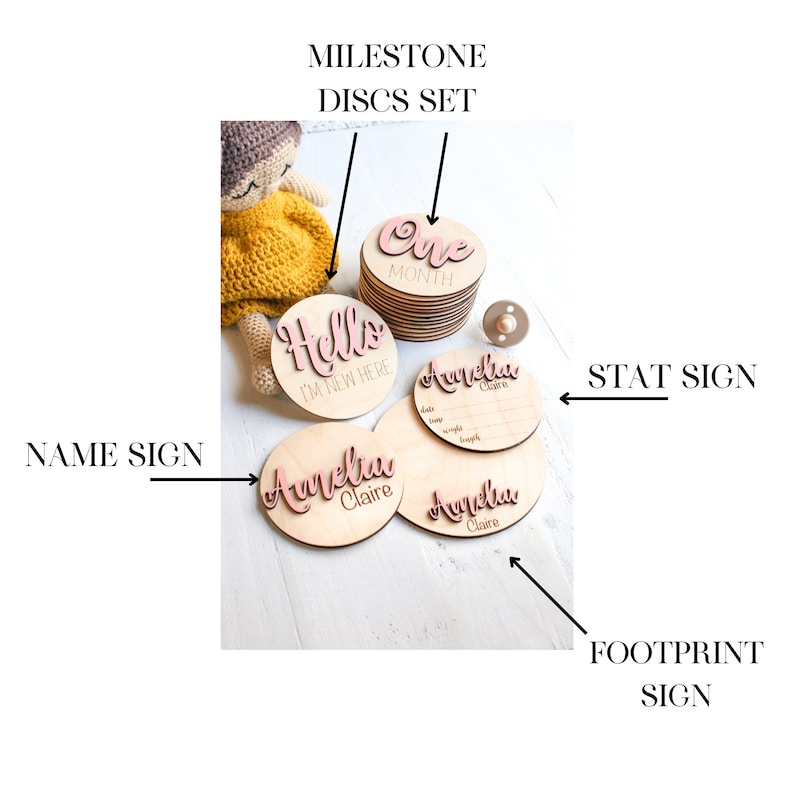 May include: Wooden baby milestone discs set with pink lettering. The set includes a "Hello I'm New Here" disc, a "One Month" disc, and three name discs that say "Amelia Claire". There is also a separate wooden stat sign with spaces for baby's name, date, time, weight, and length.
